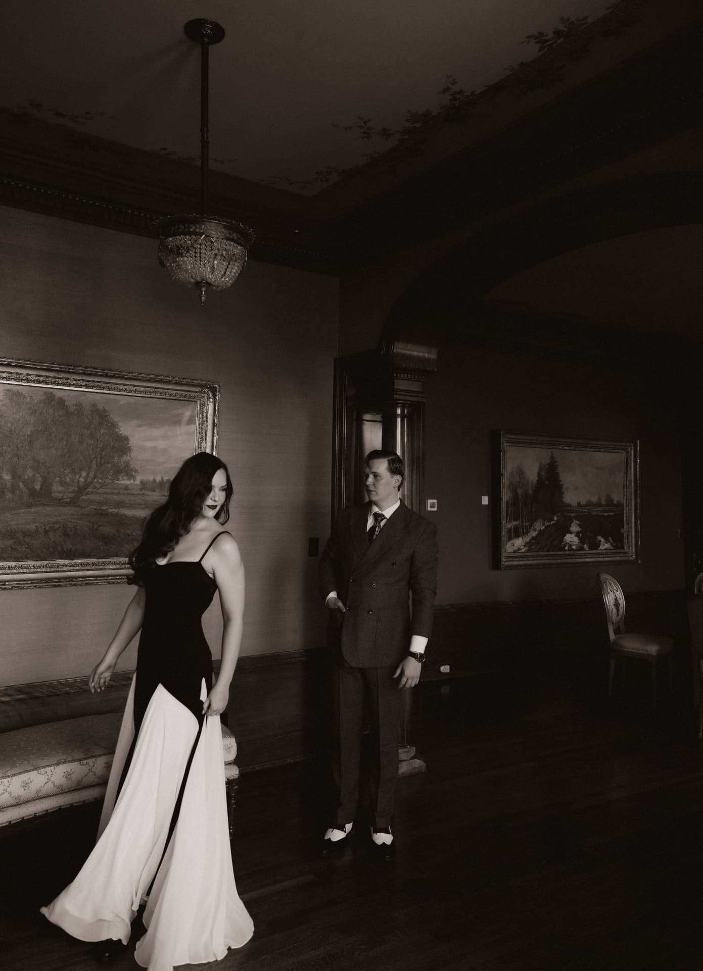 Drew and Olympia in a portrait-lined parlour, black and white